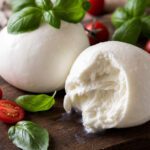 Zwei Kugeln Büffelmozzarella auf einem Holzbrett, eine davon aufgeschnitten, mit Tomaten und Basilikum