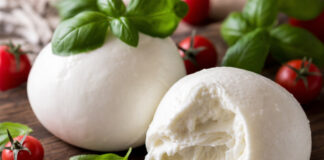 Wie schmeckt Büffelmozzarella? Zwei Kugeln Büffelmozzarella auf einem Holzbrett, eine davon aufgeschnitten, mit Tomaten und Basilikum