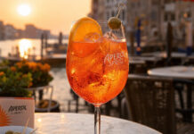 Wie schmeckt Aperol? Aperol Spritz Cocktail im Glas mit Eis und Orangenscheibe auf einer Terrasse