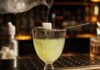 Wie schmeckt Absinth? Absinthglas mit hellgrüner Spirituose, darüber ein perforierter Löffel mit Zuckerwürfel, auf den Eiswasser aus einer Karaffe tropft