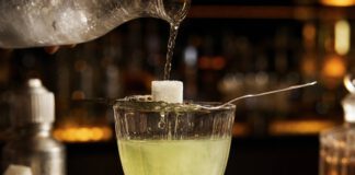Wie schmeckt Absinth? Absinthglas mit hellgrüner Spirituose, darüber ein perforierter Löffel mit Zuckerwürfel, auf den Eiswasser aus einer Karaffe tropft