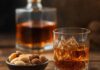 Wie schmeckt Amaretto? Glas mit bernsteinbraunem Amaretto auf Eis, davor eine Schale mit blanchierten Mandeln und Aprikosenkernen, dahinter eine eckige Likörkaraffe