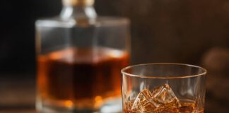 Wie schmeckt Amaretto? Glas mit bernsteinbraunem Amaretto auf Eis, davor eine Schale mit blanchierten Mandeln und Aprikosenkernen, dahinter eine eckige Likörkaraffe