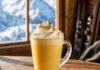 Was ist Bombardino? Glasbecher mit Henkel voll heißem gelbem Bombardino und hoher Sahnehaube mit Zimt, auf einem Holztisch vor einem Fenster mit verschneiten Bergen
