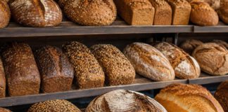 Tag des Brotes: Zwei Termine, Kulturerbe und was drin sein muss Verschiedene Brotlaibe im Holzregal einer Bäckerei, von dunklem Roggenbrot bis zu hellem Weizenbrot, ein Laib aufgeschnitten