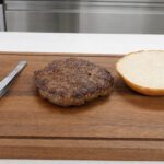 Gebratenes Burger-Patty auf einem Holzbrett neben einer Brötchenhälfte und einer Pinzette