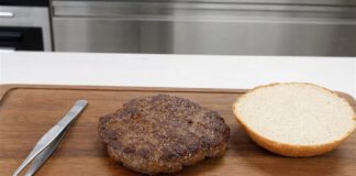 Wie schmeckt Laborfleisch? Gebratenes Burger-Patty auf einem Holzbrett neben einer Brötchenhälfte und einer Pinzette