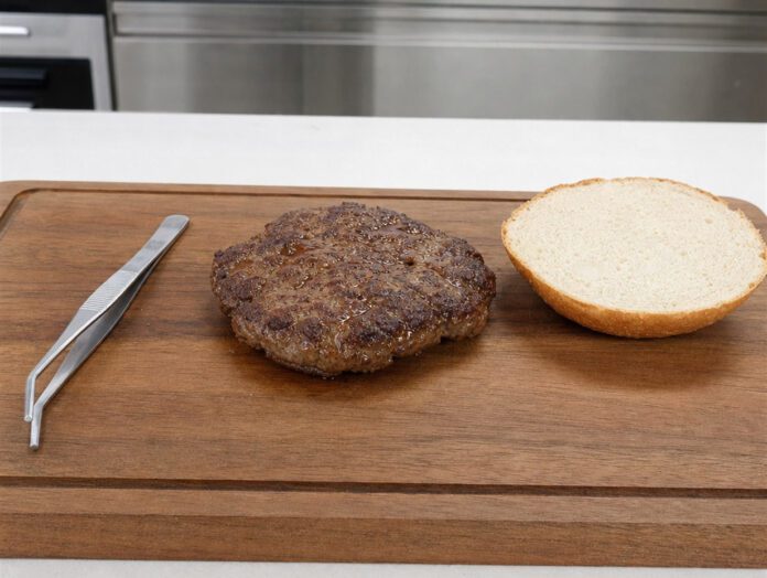 Gebratenes Burger-Patty auf einem Holzbrett neben einer Brötchenhälfte und einer Pinzette