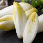 Mehrere Chicorée-Köpfe mit elfenbeinweißen Blättern und gelben Spitzen auf Schiefer, einer längs halbiert