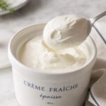 Weißes Keramiktöpfchen mit dicker glänzender Crème fraîche, ein Löffel hebt eine standfeste Portion heraus