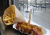 Tag der Currywurst am 4. September: Herta Heuwer und ihre Sauce Portion Currywurst in einer Papierschale mit dicker roter Currysauce, Currypulver und Holzgabel, daneben Pommes