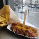 Portion Currywurst in einer Papierschale mit dicker roter Currysauce, Currypulver und Holzgabel, daneben Pommes