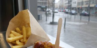 Tag der Currywurst am 4. September: Herta Heuwer und ihre Sauce Portion Currywurst in einer Papierschale mit dicker roter Currysauce, Currypulver und Holzgabel, daneben Pommes