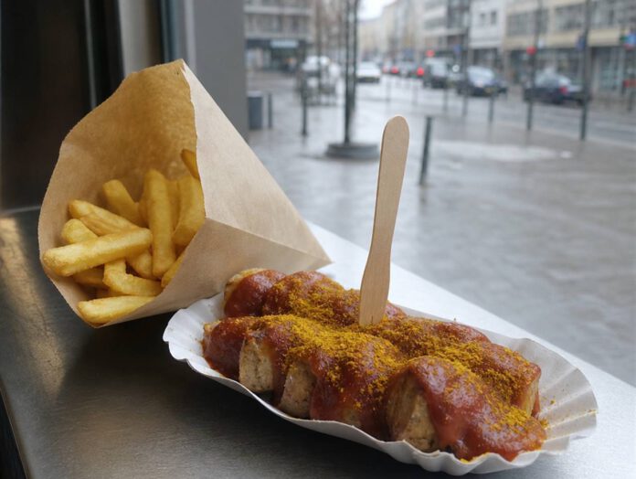 Portion Currywurst in einer Papierschale mit dicker roter Currysauce, Currypulver und Holzgabel, daneben Pommes