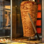 Senkrechter Dönerspieß hinter Glas an einem Imbiss, die äußere Schicht ist gebräunt, daneben ein langes Messer