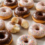 Glasierte Ringdonuts mit Zuckerstreuseln und Schokoglasur auf Backpapier, einer aufgebrochen
