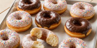 Tag des Donuts: Herkunft aus dem Krieg und der Unterschied zum Berliner Glasierte Ringdonuts mit Zuckerstreuseln und Schokoglasur auf Backpapier, einer aufgebrochen
