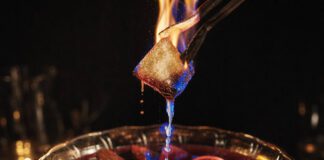 Was ist Feuerzangenbowle? Brennender Zuckerhut auf einer Metallzange über einer Bowleschale mit rotem Glühwein, Zimtstangen und Orangenscheiben, karamellisierter Zucker tropft in blauer Flamme hinunter