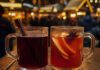 Glühwein oder Punsch: Was ist der Unterschied? Zwei Glasbecher nebeneinander auf einem Holztisch, links dunkelroter Glühwein mit Zimtstange, rechts orangeroter Fruchtpunsch mit Apfel- und Orangenscheiben und Sternanis