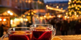 Was ist Glühwein? Zwei dampfende Glastassen mit rotem Glühwein und Orangenscheiben auf einem Holztisch, dahinter unscharfe Lichter eines Weihnachtsmarkts
