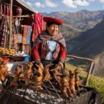 Marktstand in Peru mit Spießen über Holzkohle vor bunten Textilien und Bergkulisse
