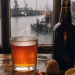 Dickwandiges Glas mit bernsteinfarbenem Grog auf einem alten Holztisch, daneben eine dunkle Rumflasche, braune Kandisstücke und eine Zitronenscheibe vor einem Hafenfenster