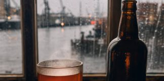 Was ist Grog? Dickwandiges Glas mit bernsteinfarbenem Grog auf einem alten Holztisch, daneben eine dunkle Rumflasche, braune Kandisstücke und eine Zitronenscheibe vor einem Hafenfenster