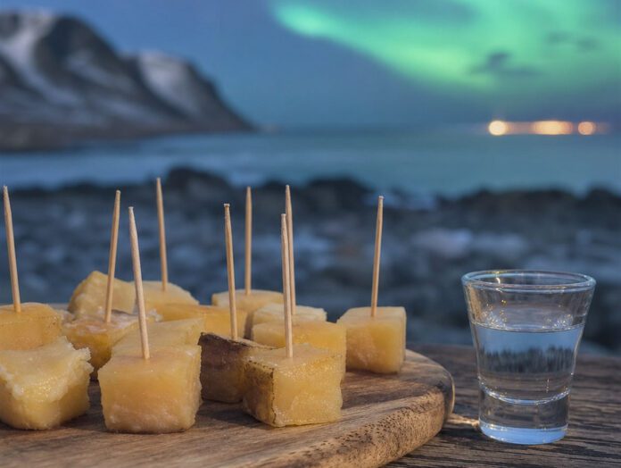Weiße Würfel fermentierten Hais auf einem Holzbrett neben einem Glas klarem Schnaps