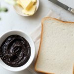Dunkler Hefeaufstrich in einer weißen Schale neben einer Scheibe Toastbrot, Butter und Messer