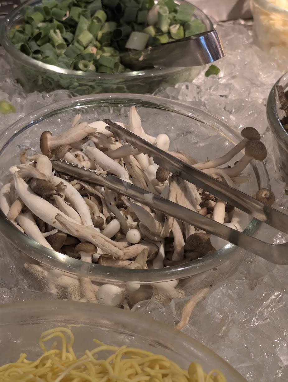 Glasschale mit Shimeji- und Austernpilzen auf Crusheis an einem Hot-Pot-Buffet, daneben Frühlingszwiebeln und Nudeln