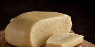 Was ist Marzipan? Angeschnittener Laib heller Marzipanrohmasse auf einem Holzbrett, davor blanchierte Mandeln und ein Häufchen Puderzucker