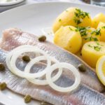 Zwei helle Matjesfilets auf weißem Teller mit rohen Zwiebelringen, Kapern, Zitrone und Salzkartoffeln