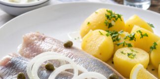 Wie schmeckt Matjes? Zwei helle Matjesfilets auf weißem Teller mit rohen Zwiebelringen, Kapern, Zitrone und Salzkartoffeln