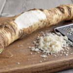 Lange knorrige Meerrettichwurzel auf einem Holzbrett, teilweise geschält, daneben frisch geriebener weißer Meerrettich und eine Reibe