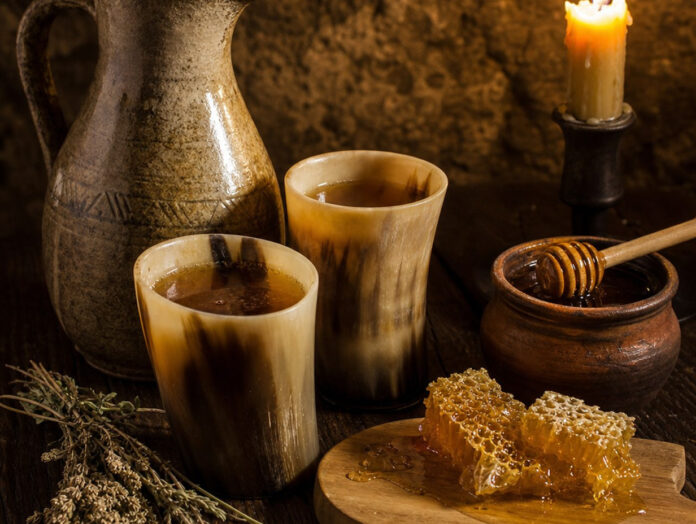 Zwei Trinkhörner mit goldgelbem Met, ein Steinzeugkrug, ein Topf Honig und eine tropfende Honigwabe auf dunklem Holz