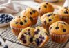 Muffin Monday: Woher der Brauch kommt und was den Muffin ausmacht Blaubeermuffins mit gewölbter Kuppe in Papierförmchen auf einem Kuchengitter, einer aufgebrochen