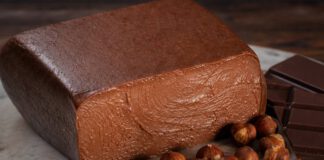 Was ist Nougat? Aufgeschnittener Block brauner Nougatmasse auf Marmor, davor geröstete Haselnüsse, blanchierte Mandeln und dunkle Schokolade