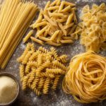 Spaghetti, Penne, Farfalle, Fusilli und ein Nest Eiernudeln auf einem bemehlten Holztisch, dazu eine Schale Hartweizengrieß