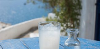 Wie schmeckt Ouzo? Hohes Glas mit milchig getrübtem Ouzo und Eiswürfeln auf einem blauen Holztisch, daneben eine Wasserkaraffe sowie Schälchen mit Feta und Oliven
