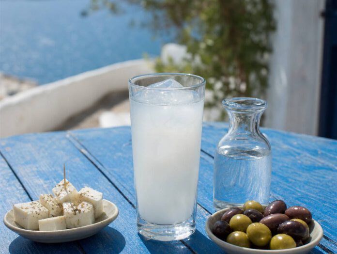 Hohes Glas mit milchig getrübtem Ouzo und Eiswürfeln auf einem blauen Holztisch, daneben eine Wasserkaraffe sowie Schälchen mit Feta und Oliven