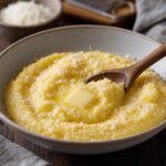 Schale mit cremiger gelber Polenta, darauf schmelzende Butter und geriebener Käse, daneben ein Holzlöffel