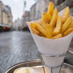 Frisch frittierte dicke Pommes frites in einer Papiertüte an einem Imbissstand, daneben ein Becher Mayonnaise