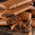 Gestapelte längliche braune Printen auf einem Holzbrett, mehrere gebrochen mit sichtbaren hellen Zuckerkristallen in der Krume, daneben brauner Kandiszucker