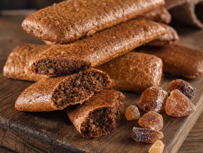 Gestapelte längliche braune Printen auf einem Holzbrett, mehrere gebrochen mit sichtbaren hellen Zuckerkristallen in der Krume, daneben brauner Kandiszucker
