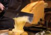 Was ist Raclette? Halber Käselaib unter einem Heizelement, die geschmolzene Schicht wird mit einem breiten Messer auf Pellkartoffeln abgeschabt, daneben Essiggurken und Silberzwiebeln