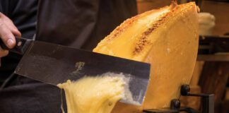 Was ist Raclette? Halber Käselaib unter einem Heizelement, die geschmolzene Schicht wird mit einem breiten Messer auf Pellkartoffeln abgeschabt, daneben Essiggurken und Silberzwiebeln