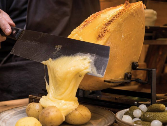 Halber Käselaib unter einem Heizelement, die geschmolzene Schicht wird mit einem breiten Messer auf Pellkartoffeln abgeschabt, daneben Essiggurken und Silberzwiebeln