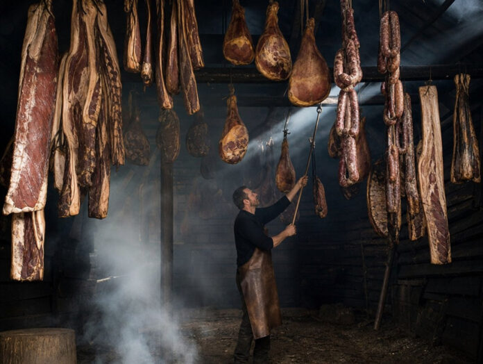 Dicht behängte Räucherkammer mit Speckseiten, Schinken und Würsten unter rußgeschwärzten Balken, im kühlen Rauch eines Schwelfeuers