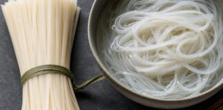 Wie schmecken Reisnudeln? Bündel trockener weißer Reisbandnudeln neben einer Schale mit eingeweichten durchscheinenden Nudeln und Stäbchen