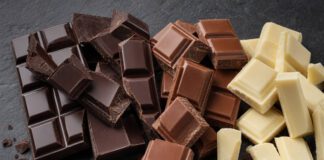 Tag der Schokolade: Welches Datum gilt und was dahintersteckt Gebrochene Stücke von Zartbitter-, Vollmilch- und weißer Schokolade nebeneinander auf Schiefer, dazu Kakaobohnen und Kakaopulver